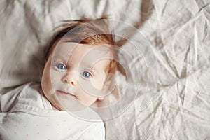 Closeup portrait of cute Caucasian red-haired newborn baby. Adorable funny child infant with blue grey eyes lying on bed looking