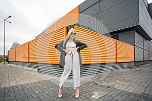 Closeup portrait of cute smiling little model girl in striped jacket and sunglasses posing near gray corrugated striped orange