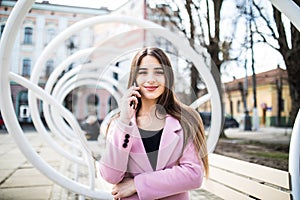 Closeup portrait of enjoyed brunette girl speaking on phone on street. She wears pink jacket, smiling to camera