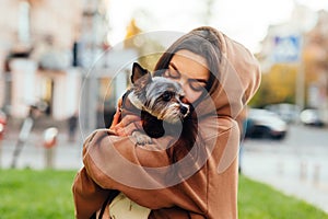 Closeup portrait of a female dog-owner wearing a hood, holding and hugging her little york terrier, feeling happy. Blurred