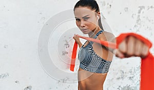 Closeup portrait of fitness woman standing against concrete wall doing stretching exercises holding a red resistance band.