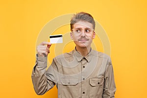 Closeup portrait of a funny nerd with a bank card in his hand on a yellow background, looking away and smiling. Guy in the shirt