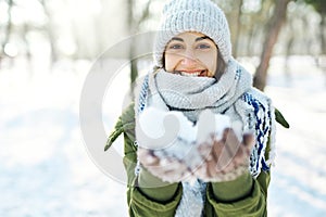 Closeup portrait of funny smiling woman in woolen hat and long warm scarf holding snow to the camera at snowy winter