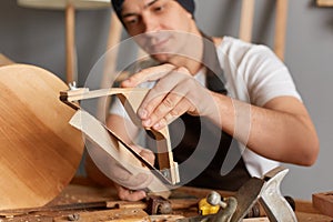 Closeup portrait of handsome young adult man carpenter wearing white T-shirt and brown apron sanding wooden wooden block, making