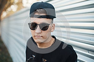 Closeup portrait of a handsome young hipster man in trendy sunglasses in a stylish black baseball cap in a shirt in the city near
