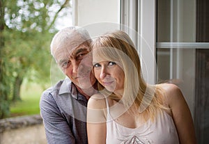 Closeup Portrait of Happy Couple with Age Difference Hugging near the Window in Their Home During Summer Hot Day