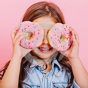 Closeup portrait happy cute little girl in blue shirt holding donuts on eyes, having fun to camera isolated on pink