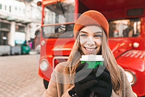 Closeup portrait of happy girl with cup of coffee standing against red bus background, looking into camera and smiling. Street