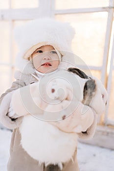 Closeup portrait of happy little child boy in warm clothes hugging white funny rabbit standing by farm house on sunny