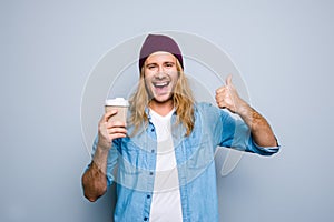 Closeup portrait of happy man gesturing his thumb finger up, holding cup of coffee smiling to the camera standing over grey