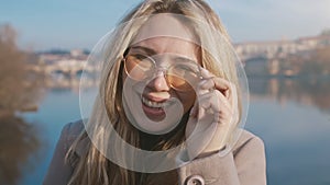 Closeup portrait of a happy young woman smiling at the camera. Natural blonde girl standing outside wearing sunglasses