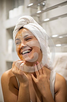 Closeup portrait laughing woman with towel on hairs posing peeling scrub mask on face at bathroom