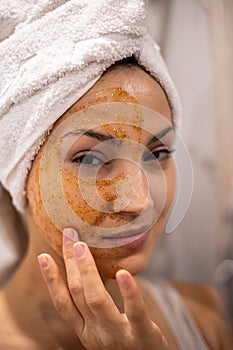 Closeup portrait laughing woman with towel on hairs posing peeling scrub mask on face at bathroom