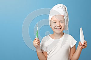 Closeup portrait of a little girl on a blue background. A child with a white towel on his head holding a toothbrush and toothpaste