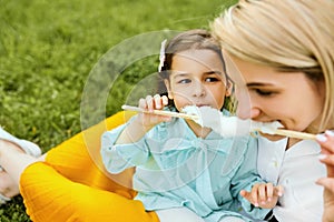 Closeup portrait of little girl eating cotton candy with her mom, sitting on the green grass in the park. Mothers day.