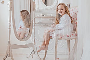 Closeup portrait of the little girl with lipstick sitting on the chair near the big mirror inside white room alone