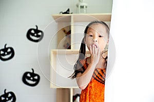 Closeup portrait little girl with surprised face in halloween costume hide behind the curtain. Decorated room in Halloween