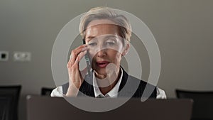 Closeup portrait of middle-aged female manager sitting at desk with laptop and talking smartphone in office