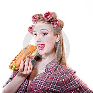 Closeup portrait of pinup beautiful blond young woman with blue eyes having fun in curlers eating hot dog on a white background