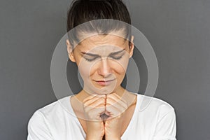 Closeup portrait of praying young female with dark hair holding her hands together at the chin, being concentrated on her wish.
