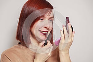 Closeup portrait redhead beautiful girl looking in mirror compact powder and paints lips with dark burgundy purple liquid lipstick