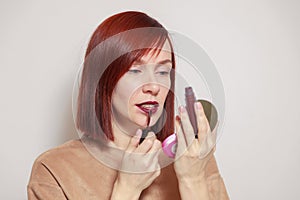 Closeup portrait redhead beautiful girl looking in mirror compact powder and paints lips with dark burgundy purple liquid lipstick