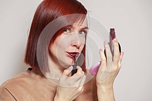 Closeup portrait redhead beautiful girl looking in mirror compact powder and paints lips with dark burgundy purple liquid lipstick