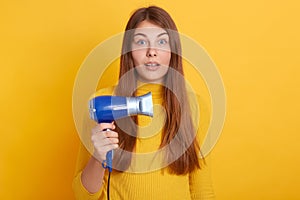 Closeup portrait of shocked female with long beautiful hair, holding hair dryer in hands, looking at camera with big eyes, being