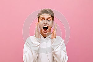 Closeup portrait of shocked guy in white hoodie on pink background looking into camera with mouth open and shouting with joy.