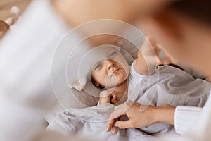 Closeup portrait of sick little baby girl with fever lying in bed under blanket, mothers hand caring for her, small female kid