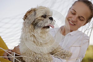 Closeup portrait of smiling beautiful cute Caucasian woman wearing white shirt lies in a hammock with Pekingese dog in nature,