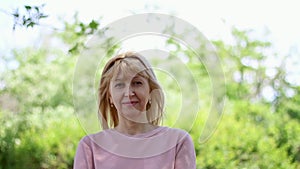 Closeup portrait of a smiling blonde middle-aged woman, she is in the park, smiling and posing on a green lilac background