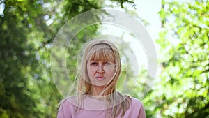 Closeup portrait of a smiling blonde middle-aged woman, she is in the park, smiling and posing on a green lilac background