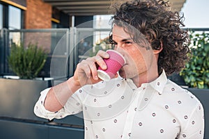 Closeup portrait of smiling Caucasian business male with curly hair and trendy shirt drinking delicious coffee and waiting for