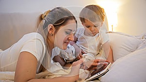 Closeup portrait of smiling young mother browsing internet on digital tablet with her baby boy in bed at night