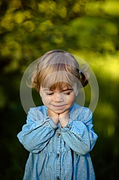 Closeup portrait tiny girl in blue jeans dress, with closed eyes, smiling to camera, on sunny green meadow. Summer time, Sunshine