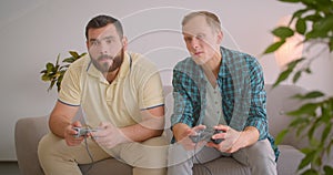 Closeup portrait of two adult men playing video games together sitting on couch at home indoors. Cheerful man winning