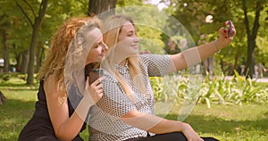 Closeup portrait of two female friends taking selfie on phone smiling happily sitting on blanket in park outdoors