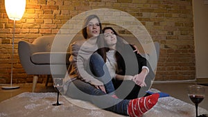 Closeup portrait of two young pretty women hugging and watching TV chilling with wine in a cozy apartment indoors