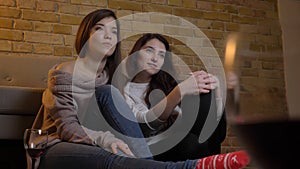 Closeup portrait of two young pretty women watching TV chilling with wine sitting on the floor in a cozy apartment