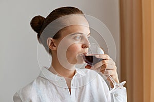 Closeup portrait of woman with bun hairstyle wearing white shirt drinking red wine from glass, looking away with dreamy expression