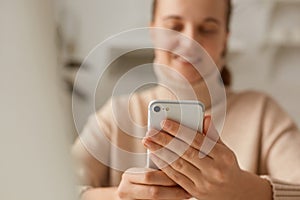 Closeup portrait of woman wearing beige sweater posing with cell phone in hands, looking smiling at devise display, communicating