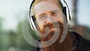 Closeup portrait of young bearded hipster man with headphones listen to music and smiling at city street