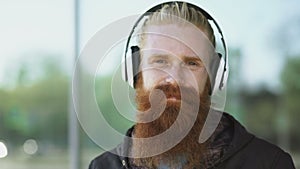 Closeup portrait of young bearded hipster man with headphones listen to music and smiling at city street