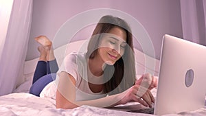 Closeup portrait of young beautiful teenage girl texting on the laptop and smiling while lying on the bed