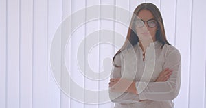 Closeup portrait of young caucasian woman in glasses looking at camera with her arms crossed over chest standing indoors