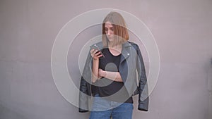 Closeup portrait of young cute caucasian girl in a leather jacket using the phone looking at camera outdoors in the