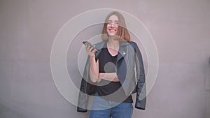 Closeup portrait of young cute caucasian girl in a leather jacket using the phone smiling happily looking at camera