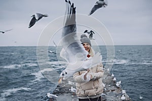 Closeup portrait of young happy woman feeds seagulls on the sea. Pretty female smiling and watching flying seagulls by the sea in