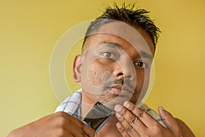 A closeup portrait of a young Indian man trimming his beard with an electric trimmer. Selective focus on beard